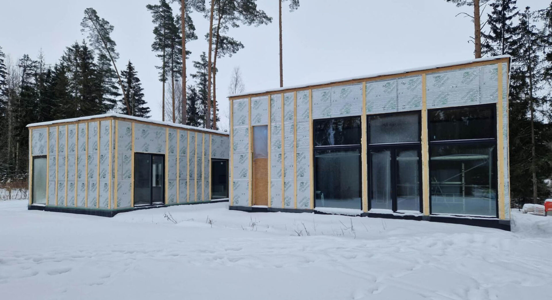 Two wings of the main house under construction in snow, insulated walls and large black-framed windows already installed.