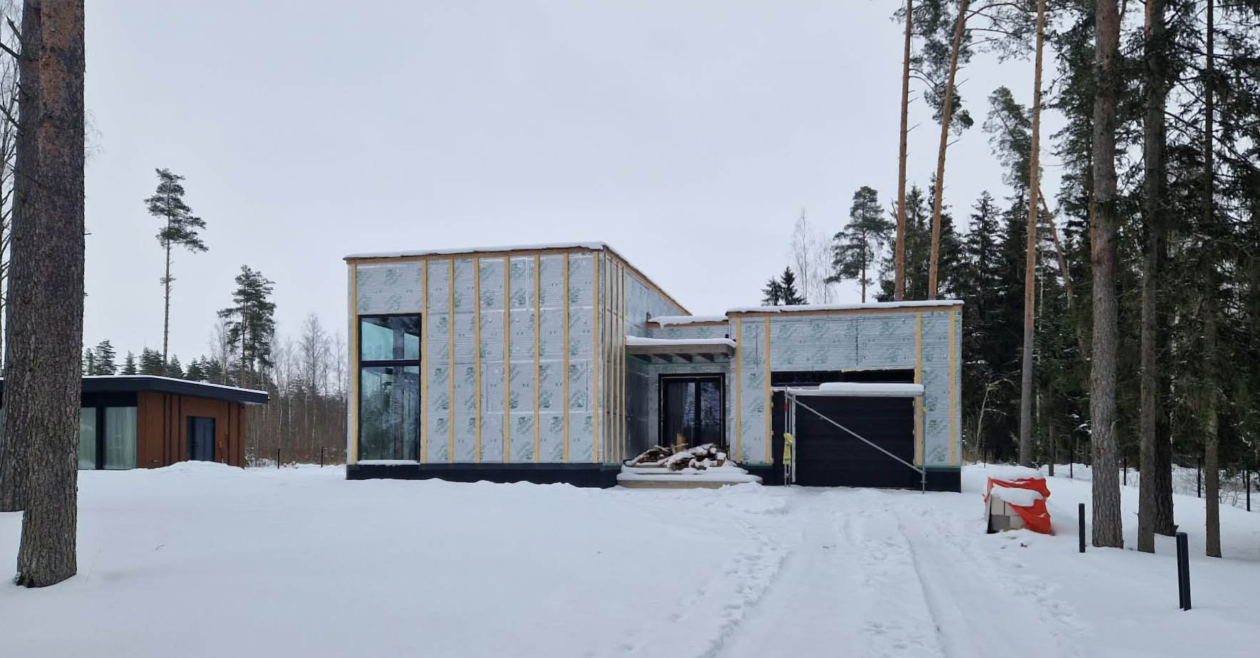 The main house under construction in winter, wrapped in exterior insulation boards, with the finished dark-clad ADU on the left.