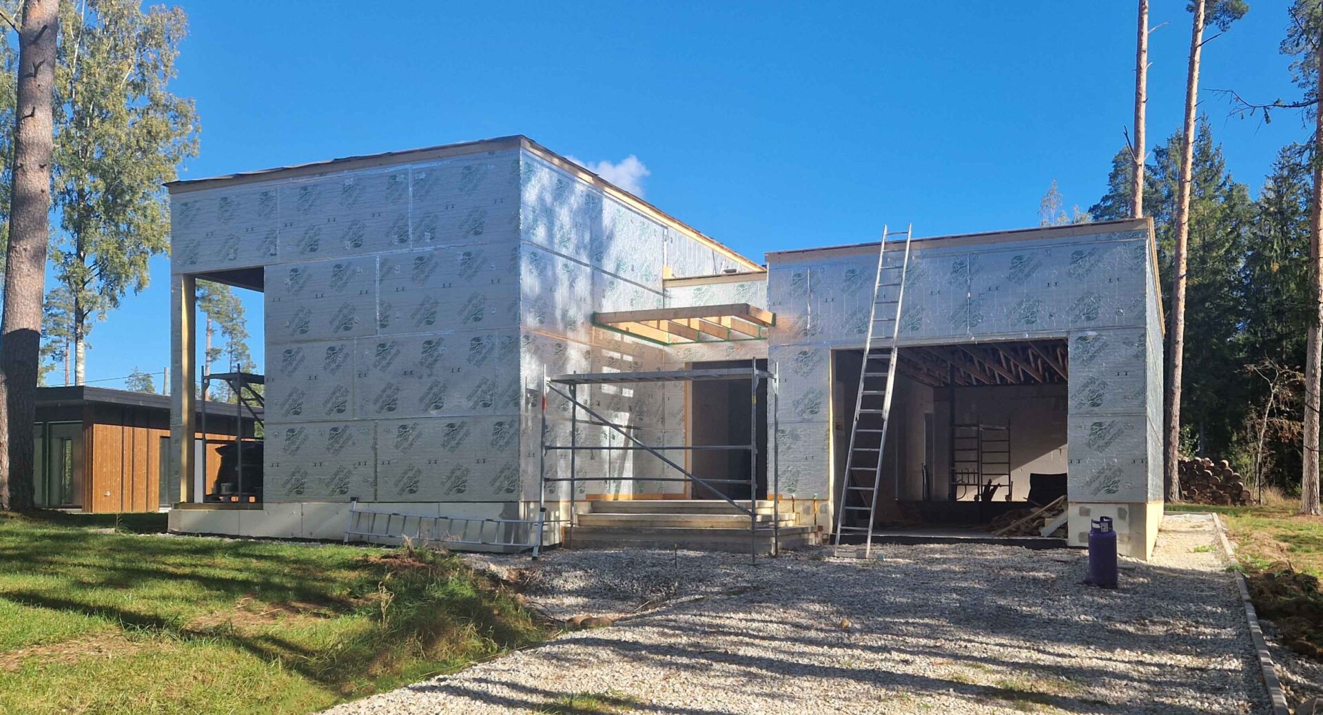 The main house in fall 2025, wrapped in exterior insulation with scaffolding at the entrance and the finished ADU on the left.