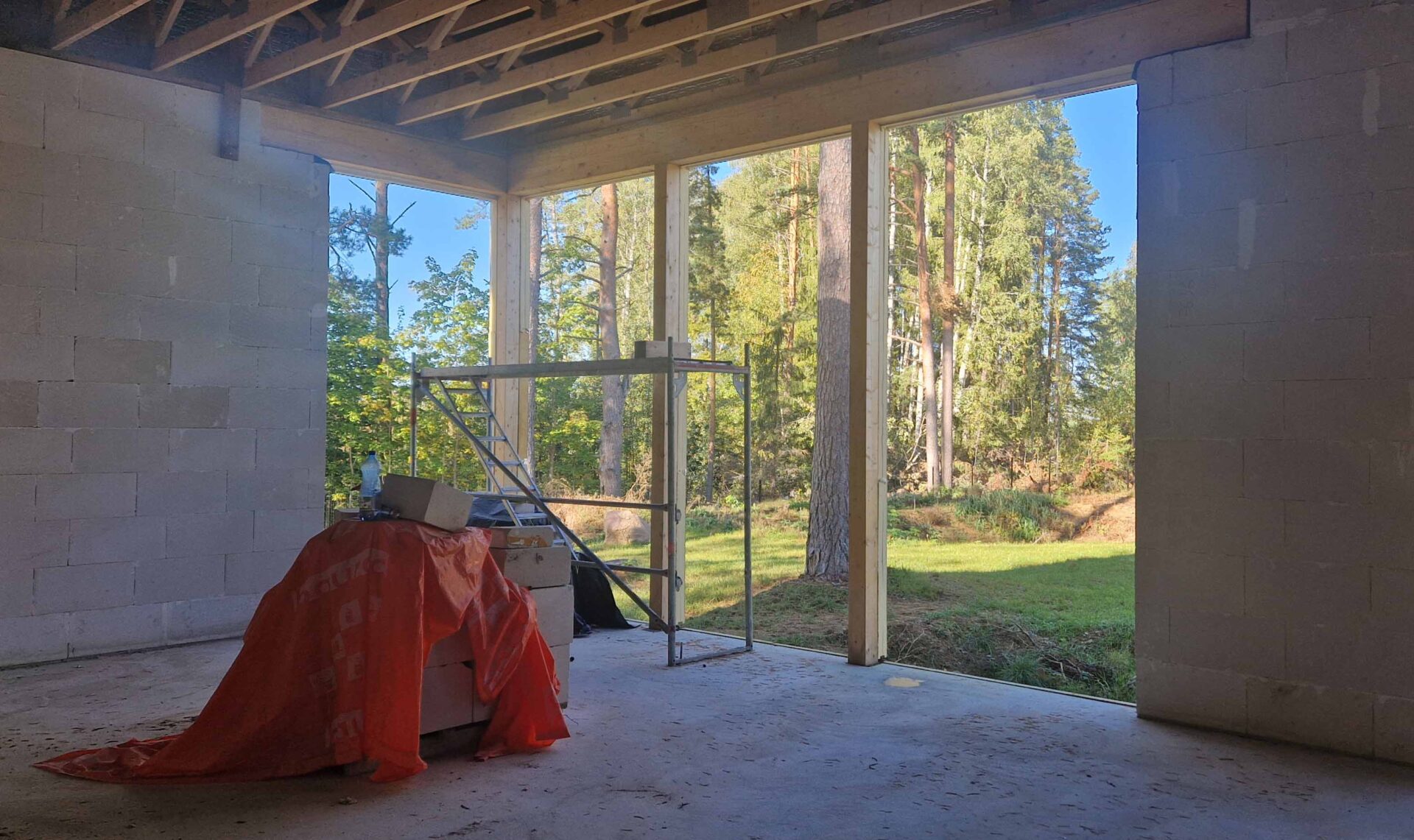 Living room under construction in fall 2025, with block walls, timber roof structure and an opening toward the pine forest.