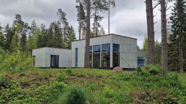 The SOLARIS house under construction in a pine forest: single-story flat-roof volumes wrapped in exterior insulation boards, with the large windows already installed.
